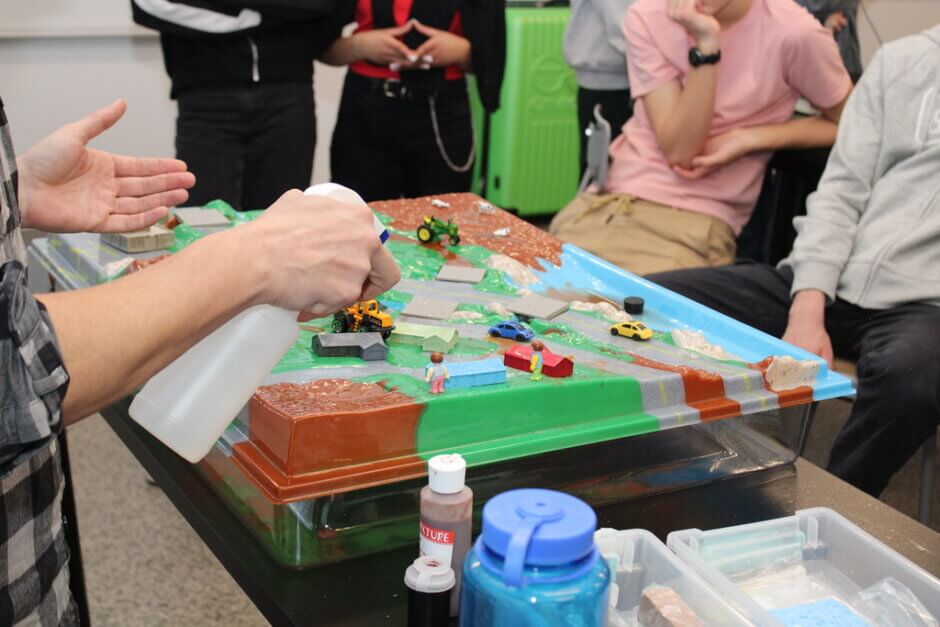 Gros plan sur la maquette du bassin versant, que l'on voit recouverte de divers bâtiments et voitures jouets. Des élèves regardent à l'arrière-plan tandis qu'une personne au premier plan explique et tient un flacon pulvérisateur pour simuler la pluie.