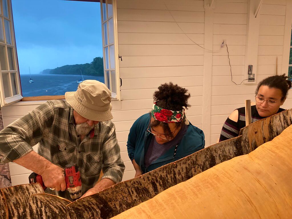 Pinock Smith travaille sur une grande section de bouleau sous la surveillance de deux jeunes participants. L'arrière-plan montre une vue de la rivière des Outaouais par la fenêtre.