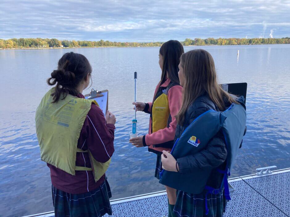 Trois étudiants portant des gilets de sauvetage se tiennent sur le pont de la Maison riveraine. L'un d'eux analyse l'eau à l'aide d'un bâton d'échantillonnage et d'un conductivimètre, tandis qu'un autre tient un presse-papiers contenant des données.