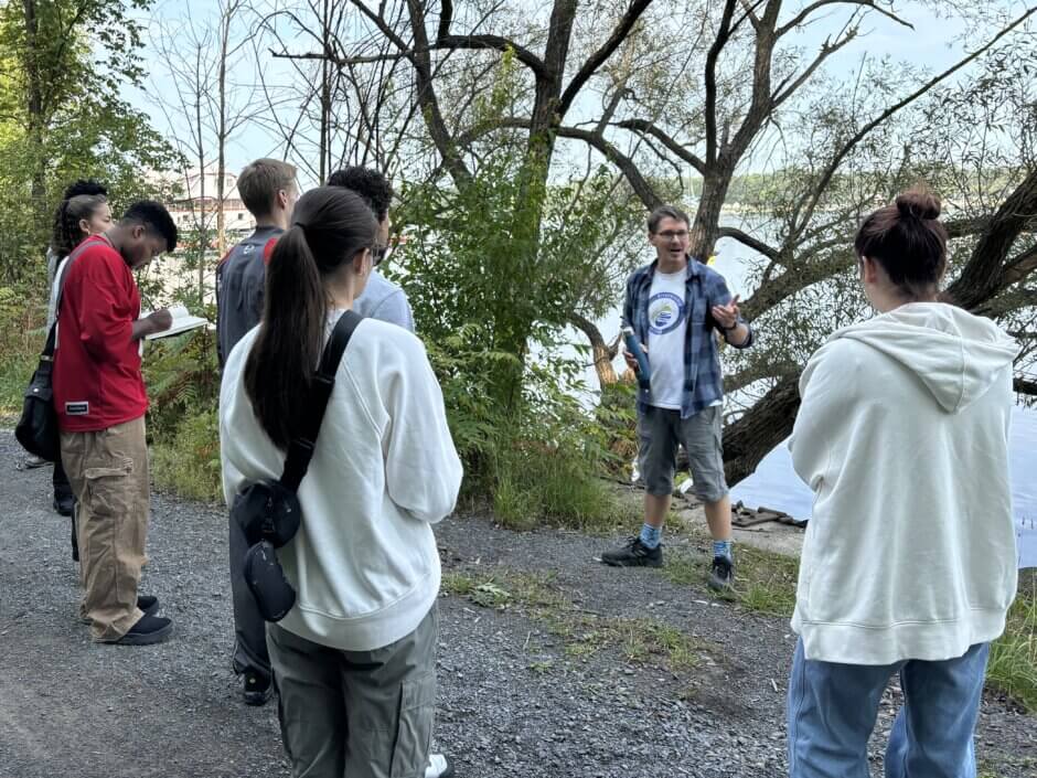 Des jeunes se tiennent à l'extérieur, le long des berges de la rivière, pour écouter une présentation.