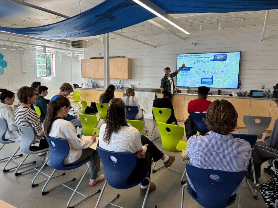 Un atelier de l'École de la rivière, montrant des étudiants en rangées de chaises regardant un éducateur pointant une carte du bassin versant de la rivière des Outaouais sur un grand écran dans le laboratoire éducatif de la Garde-rivière des Outaouais.