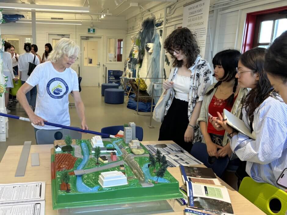Un bénévole vêtu d'une chemise de Garde-rivière des Outaouais montre à quatre jeunes le maquette du bassin versant sur une table du Labo éducatif.